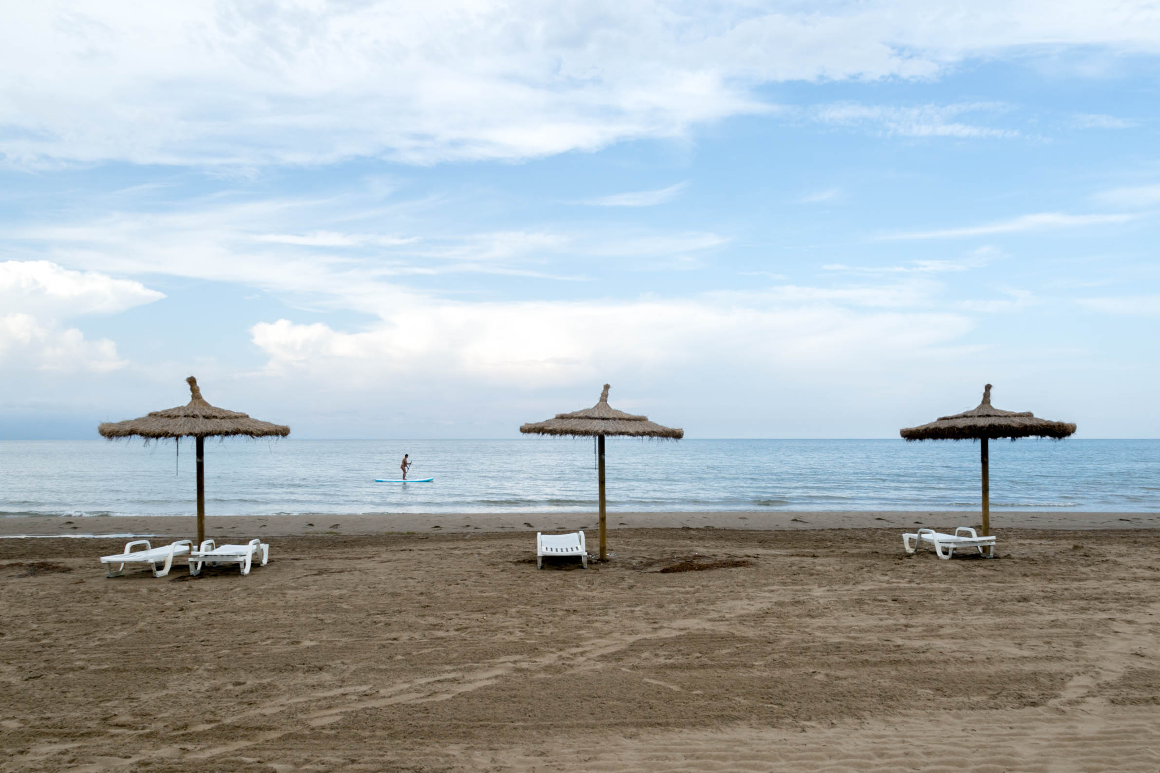 Out of season: empty beach with three parasols made of a wooden pole and straw and four white plastic sun loungers. Close to the shoreline is a woman rowing by on a stand-up paddleboard.