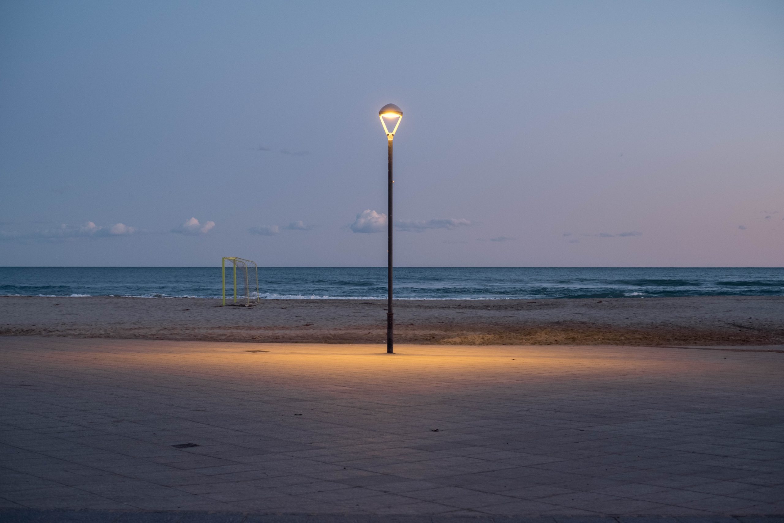 A street light set against the open horizon and the sea, projects its light onto the pavement. Behind it, on the beach is a small yellow goal.